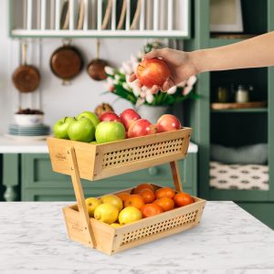 2 tier bamboo fruit basket holding apples and citrus as a hand removes an apple from the upper basket