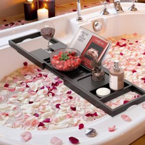 Black bamboo bathtub tray spanning a bathtub with a book, fruit, stemmed glass and toiletries