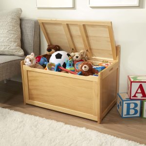 Wooden toy chest with a raised lid holding stuffed toys and a ball beside a sofa