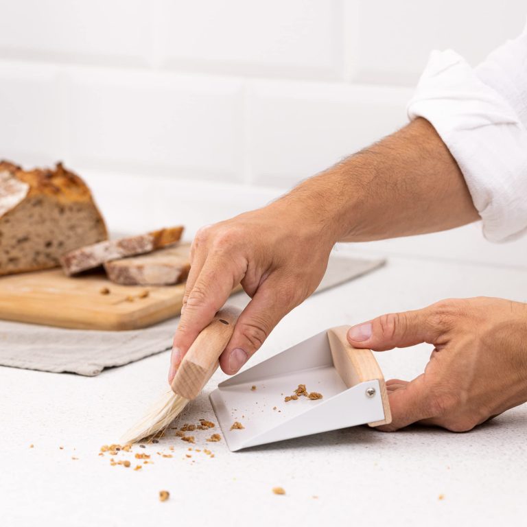 Hands using a wooden dustpan and brush set to sweep bread crumbs from a kitchen counter