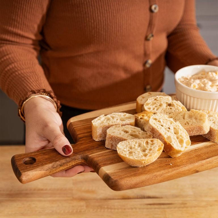Person holding the long handle of a wood cutting board arranged with sliced bread and a bowl of dip