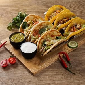 Wooden taco holder serving tray pictured with tacos in two metal racks and two black dip bowls