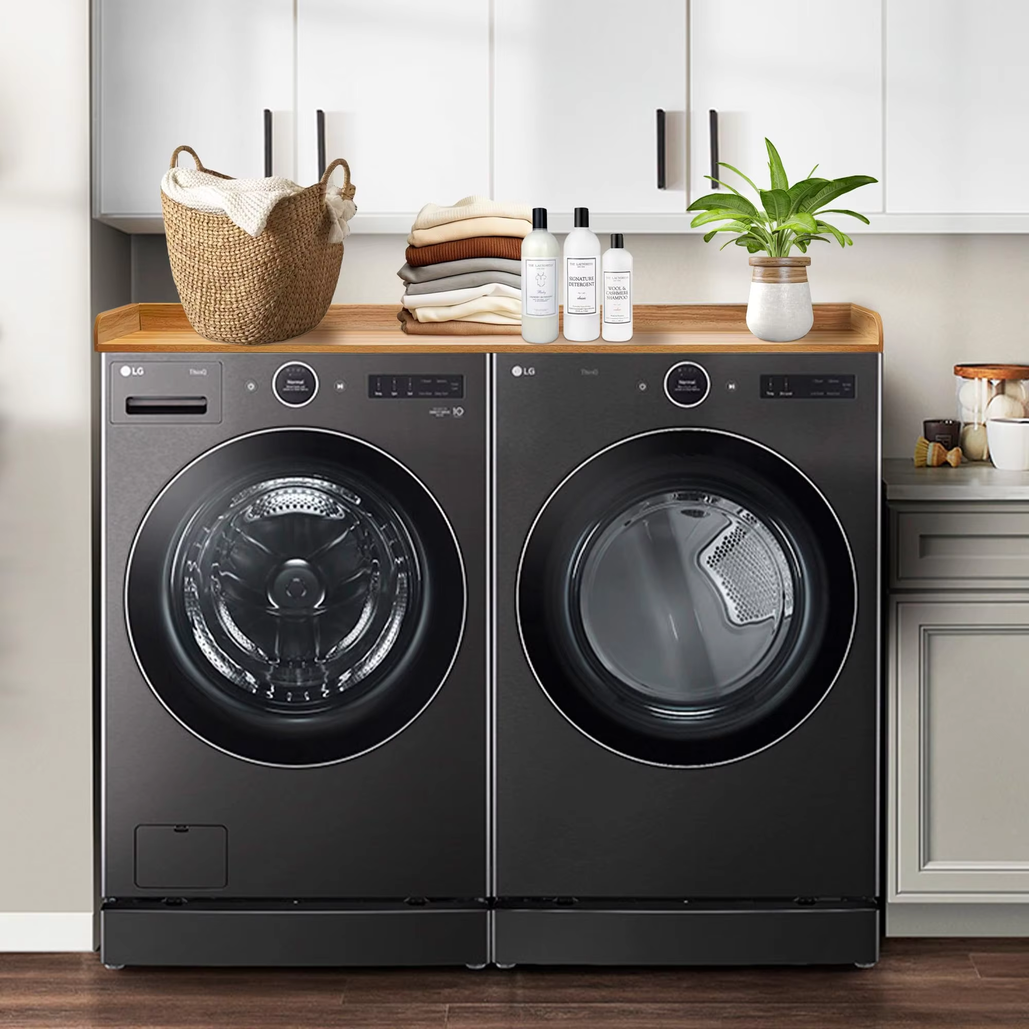 Laundry countertop above two dark front-loading appliances, pictured with folded clothes, a basket, bottles and a plant