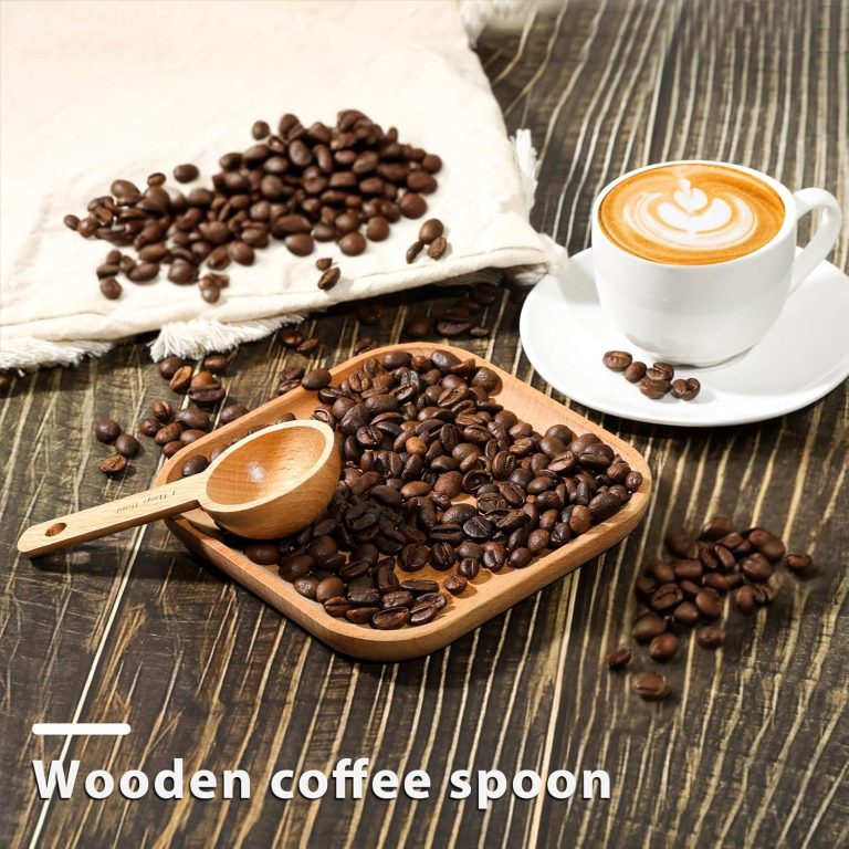 Wooden coffee measuring spoon resting on a tray of roasted beans beside a cup of coffee