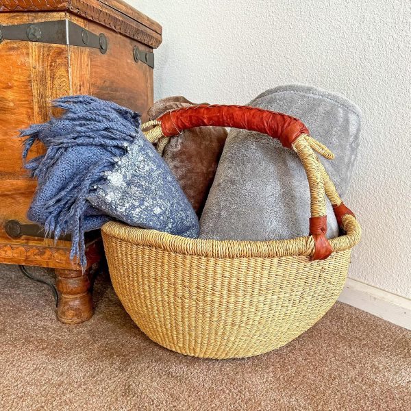 Round natural grass basket with a leather handle holding folded blankets beside a wooden chest