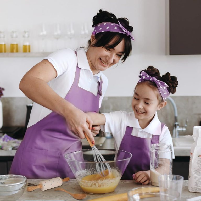 An adult guides a child using a wooden-handled whisk, with a small rolling pin and spoon on the counter