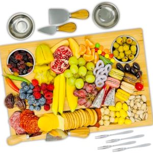 Bamboo cheese board with knives, bowls and forks arranged with cheese, fruit, crackers and small snacks