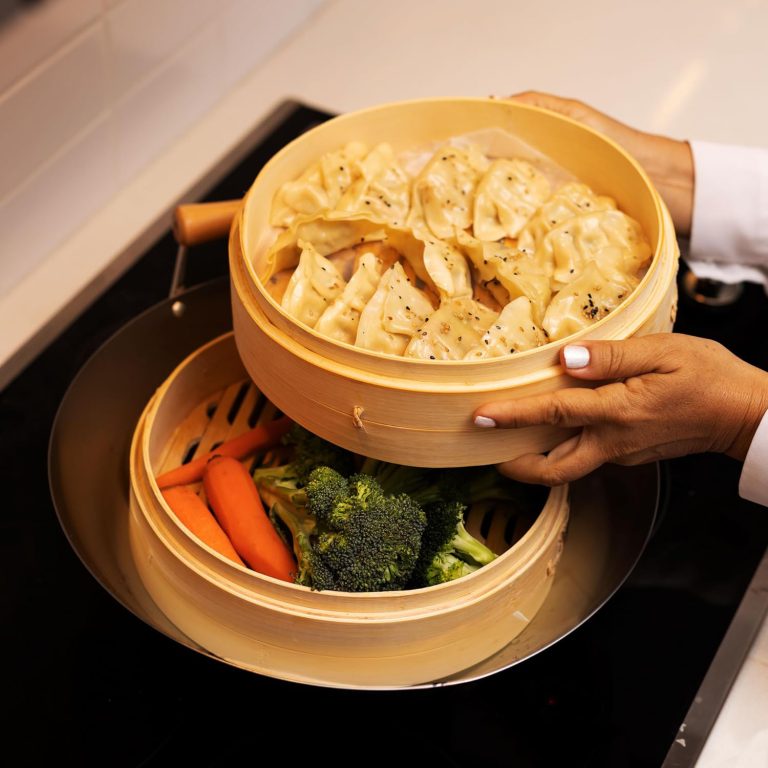 Upper bamboo steamer basket with dumplings lifted above a lower basket containing vegetables