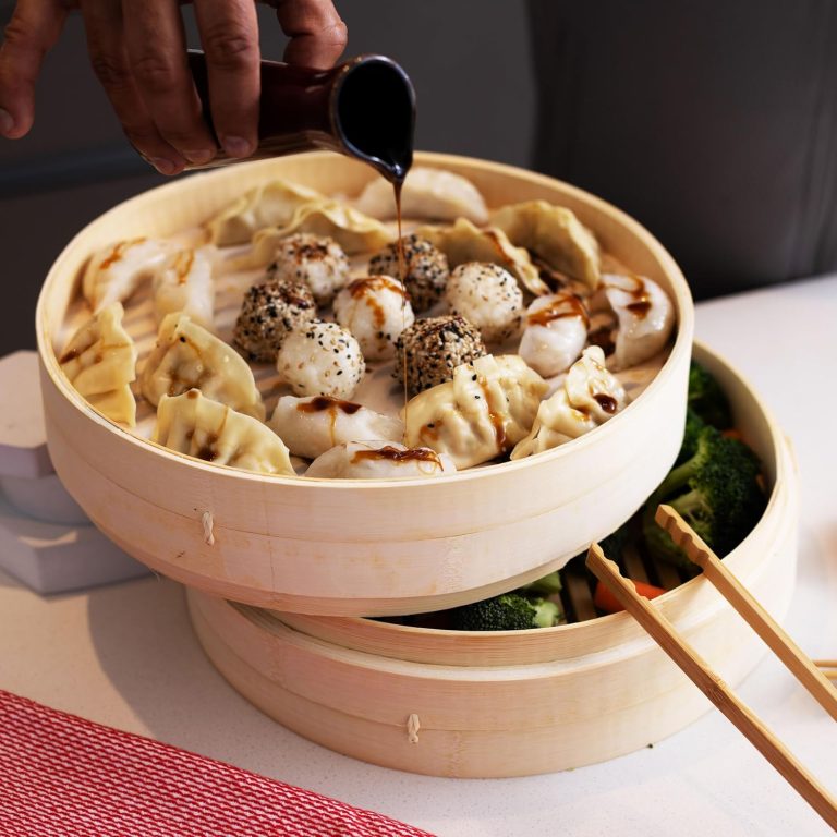 Bamboo steamer baskets with dumplings and vegetables beside a pair of serving tongs