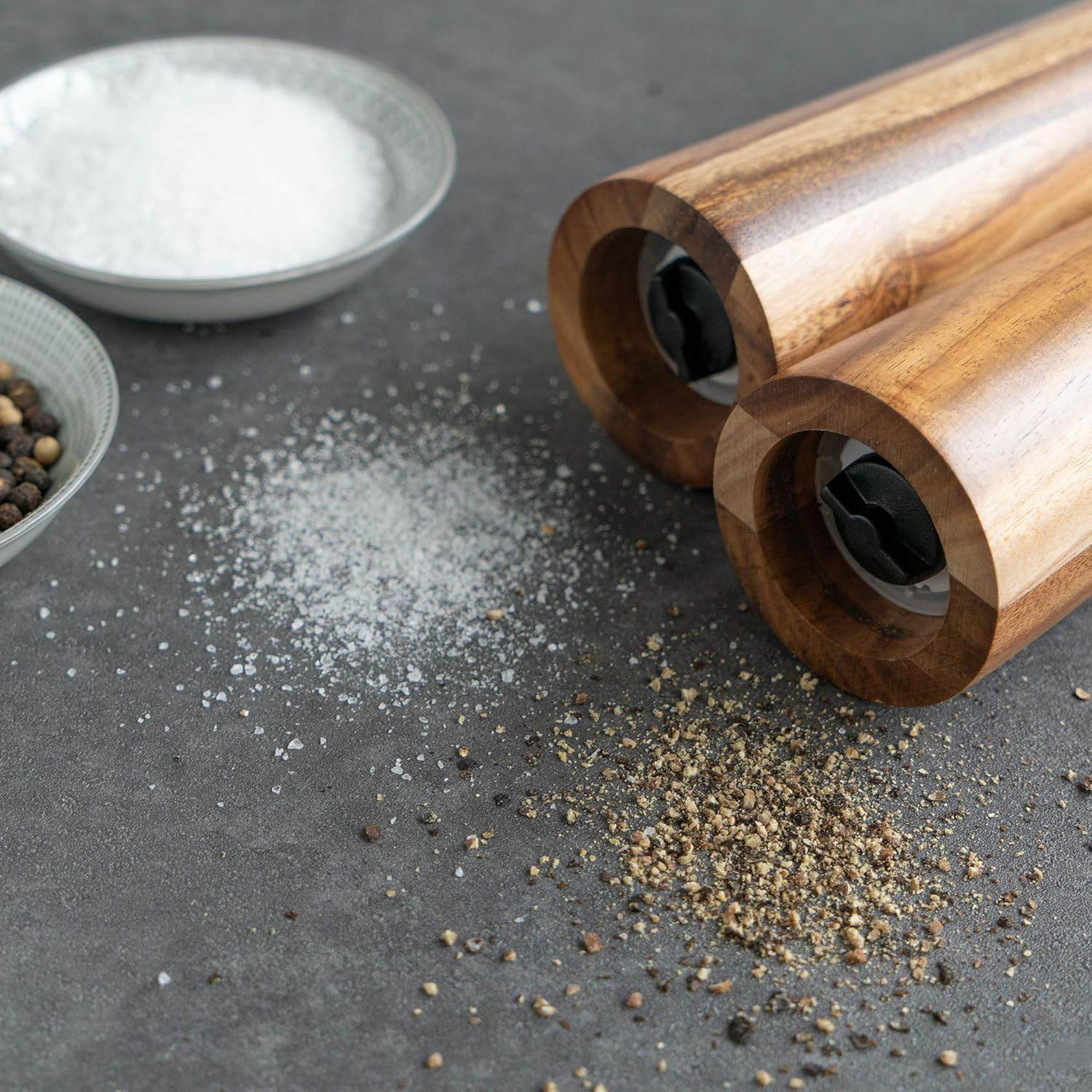 Close view of two acacia wood mill bases beside bowls of salt and peppercorns and freshly ground seasoning