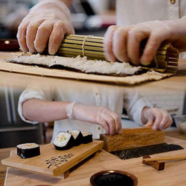 Hands demonstrating a bamboo sushi rolling mat and a wooden maki press beside a floral serving plate